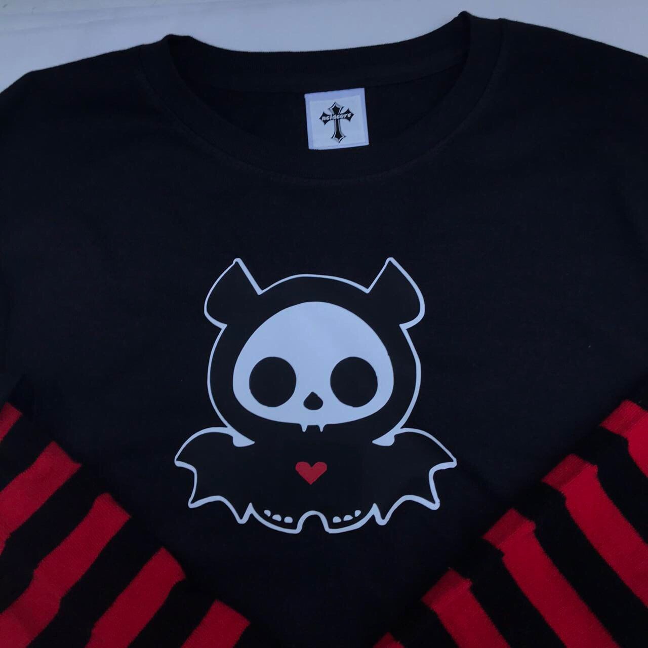 Black long-sleeved shirt with red and black striped sleeves. The shirt features a white graphic of a cartoon bat with a skull face and a red heart.